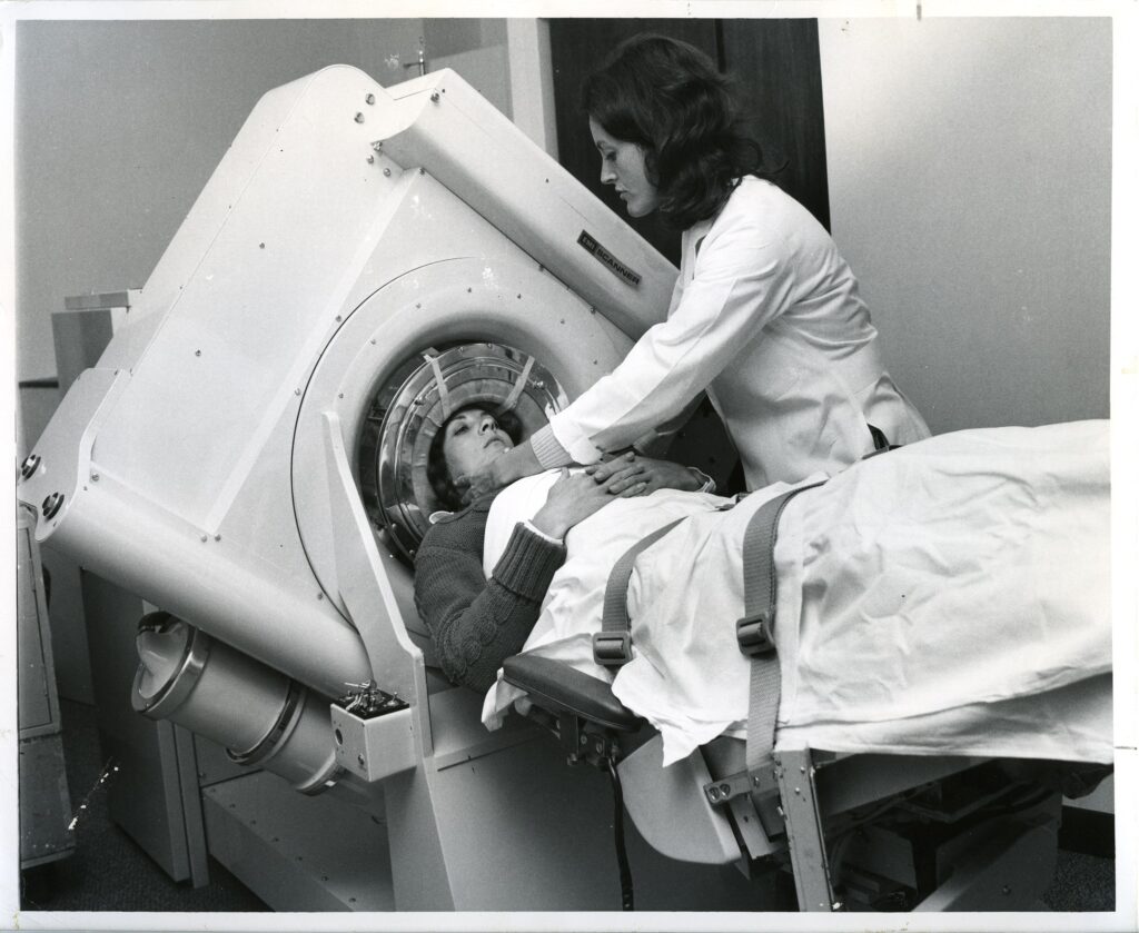 EMI Scanner; verso: "The EMI scanner is an example of recent technological advances that reduces patient X-ray exposure while providing more accurate diagnosis than obtainable with older X-ray machines. Here technician Mary McNally and secretary Penny Foster, as patient, demonstrate the giant 2 1/2 ton machine which diagnoses brain disorders." Photograph by Juan Saenz. Photograph collection, R, Radiology CT and EMI scanning.