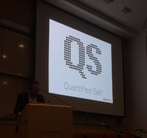 A slide shows in a darkened room as a person gives a presentation on "QS" or Quantified Self.