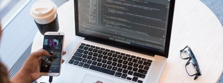 Person working on a laptop with code on the screen, holding a smartphone. A coffee cup and glasses are on the table.